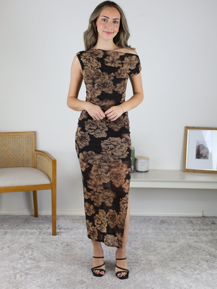 A woman wears the Espresso Garden Maxi Dress, a sleeveless, form-fitting metallic floral maxi dress in a brown and black pattern with a side slit, paired with black strappy heels.