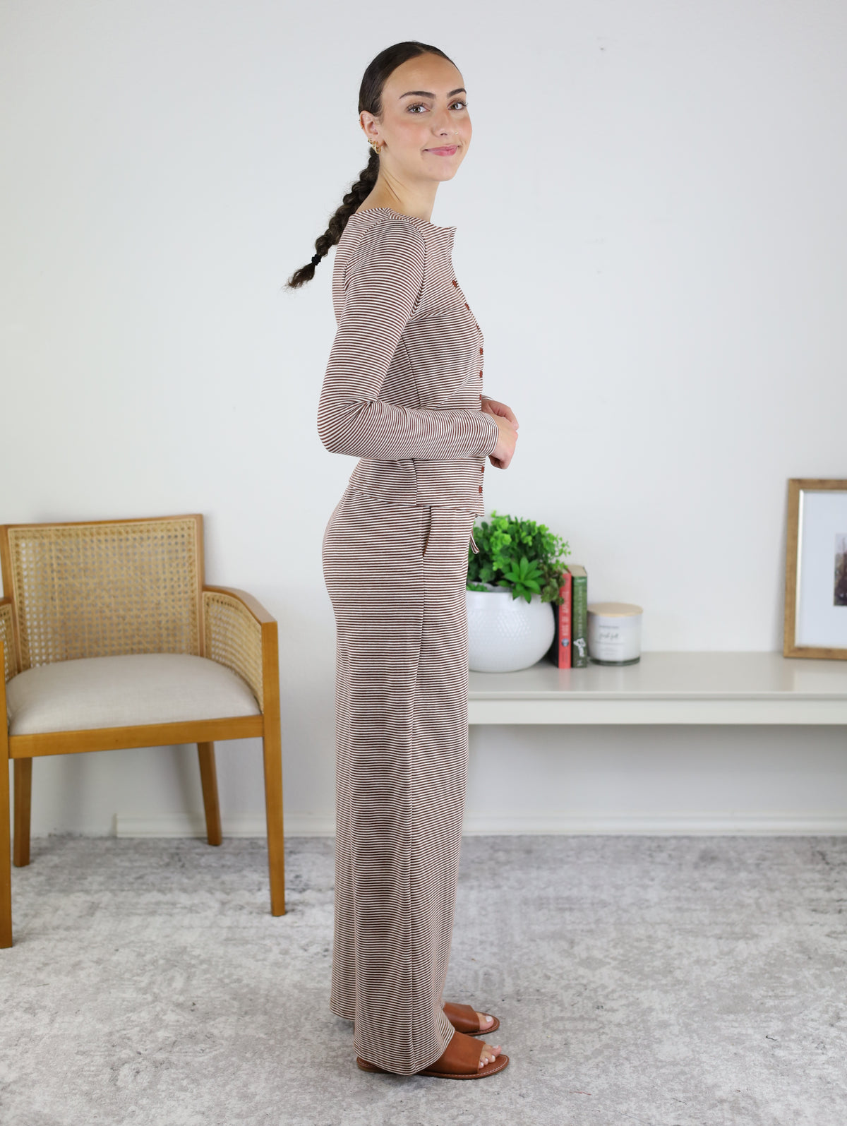 A woman with a braid stands sideways, wearing the Melissa Lounge Stripe Button Down Set, featuring a taupe long-sleeve top and matching wide-leg pants. She is indoors, near a wooden chair, a white shelf with books, a plant, and framed art.