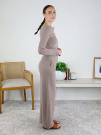 A woman with a braid stands sideways, wearing the Melissa Lounge Stripe Button Down Set, featuring a taupe long-sleeve top and matching wide-leg pants. She is indoors, near a wooden chair, a white shelf with books, a plant, and framed art.