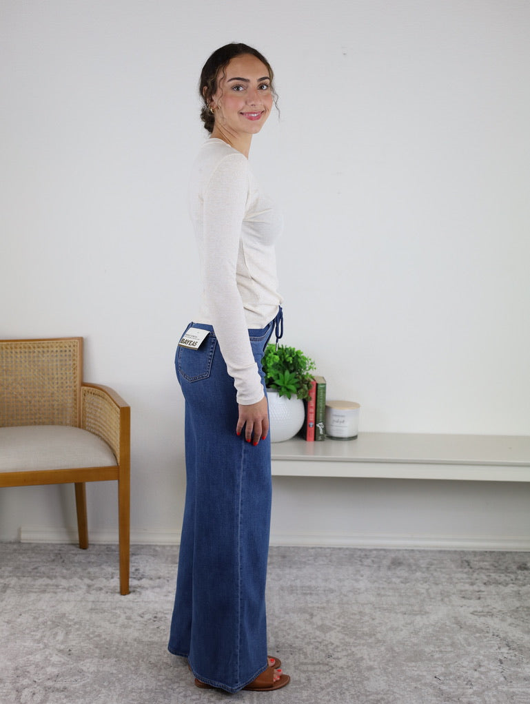 A woman stands sideways indoors wearing the Josette Ribbed Classic Crewneck—a white, long sleeve top crafted from a soft modal-spandex blend—paired with blue wide-leg jeans and brown sandals. Behind her are a wooden chair and a console table topped with plants, books, and a candle.