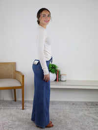 A woman stands sideways indoors wearing the Josette Ribbed Classic Crewneck—a white, long sleeve top crafted from a soft modal-spandex blend—paired with blue wide-leg jeans and brown sandals. Behind her are a wooden chair and a console table topped with plants, books, and a candle.