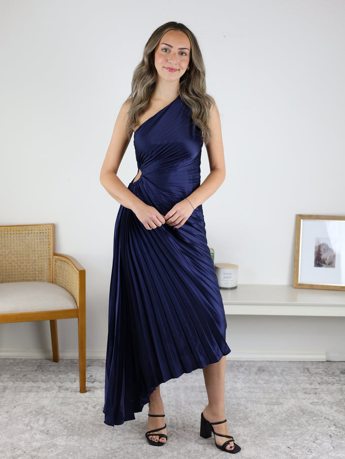 A woman wearing a dark blue Midi Satin One Shoulder Asymmetrical Pleated Dress with a one-shoulder neckline, pleated fabric, an asymmetrical hem, and a side cutout.
