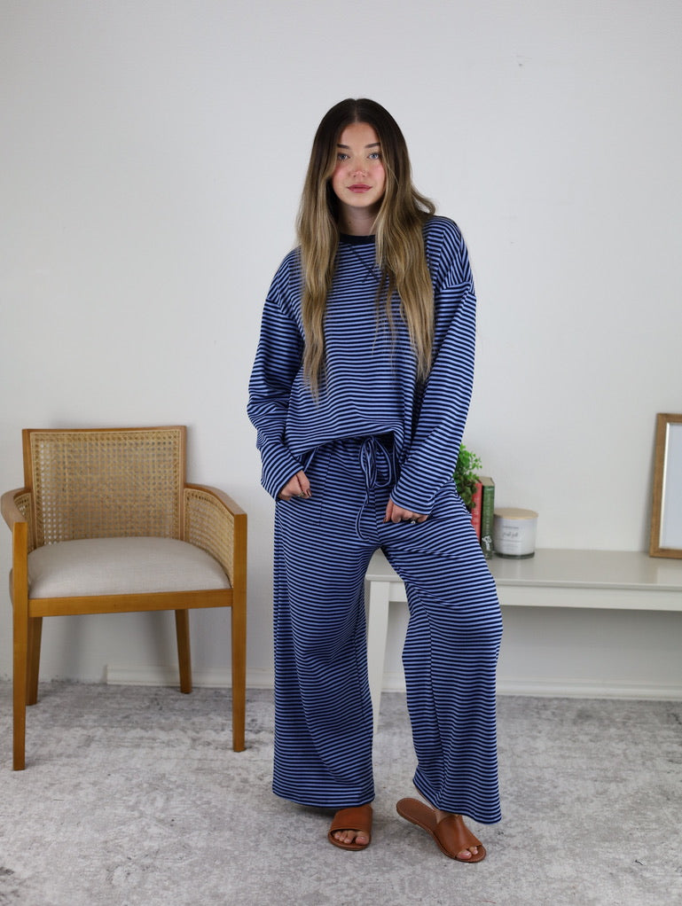 A woman stands indoors on a gray rug wearing the Misty Striped Relaxed Top & Wide Leg Pants with brown sandals. Behind her are a woven chair, a console table with decor, and a blank wall.