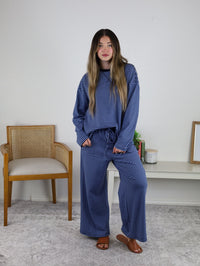 A woman stands indoors on a gray rug wearing the Misty Striped Relaxed Top & Wide Leg Pants with brown sandals. Behind her are a woven chair, a console table with decor, and a blank wall.