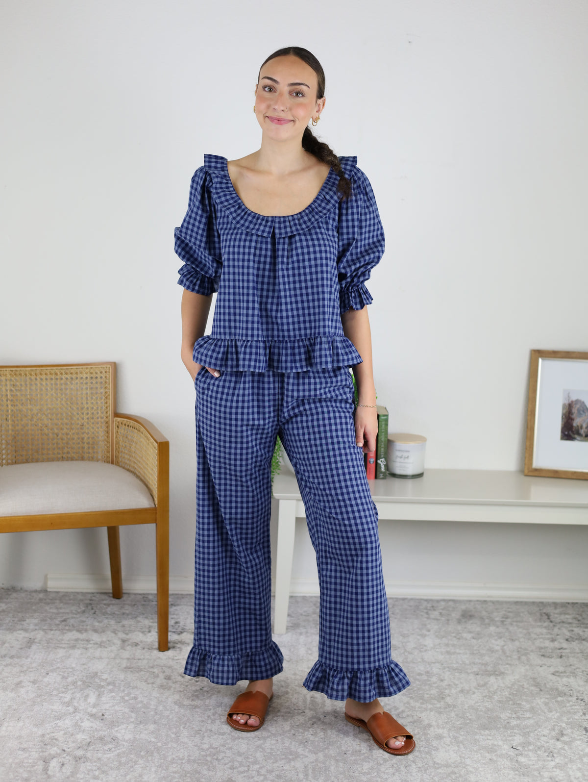 A woman stands indoors wearing the Barron Cotton Gingham Frill Set—a matching blue cotton ensemble featuring a frill top and pants with ruffle details—paired with brown sandals and her hair tied back. She stands near a wooden chair and a white shelf adorned with decor, effortlessly embodying women’s loungewear style.