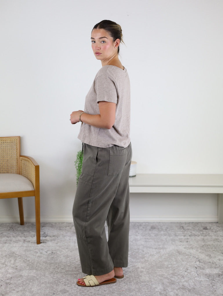 A woman stands indoors on a gray rug, wearing the Nomad V Neck Tee in a soft beige knit, paired with olive green pants and cream sandals. She faces sideways, looking at the camera. A wooden chair and a white bench are visible in the background.