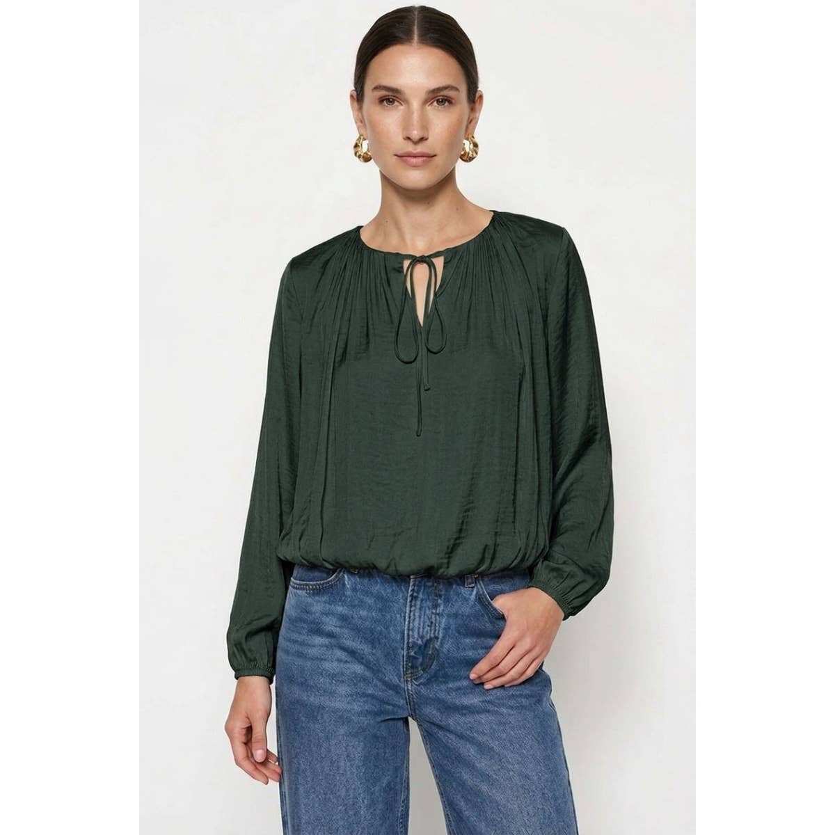 A woman wears a Naked Zebra - Long Sleeve Draped Bubble Hem Blouse: Wine / L, featuring a dark green satin fabric with a tied neckline and gathered bubble hem, paired with blue jeans.