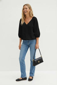 A smiling woman wears a Naked Zebra - Elastic Puff Sleeve V-neck Blouse: Burgundy / L, blue jeans, and black ballet flats, while carrying a black shoulder bag.