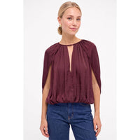 A woman wearing a dark red, silky Naked Zebra - Silky Draped Cape Sleeve Blouse: Wine / L with a keyhole neckline, paired with blue jeans.