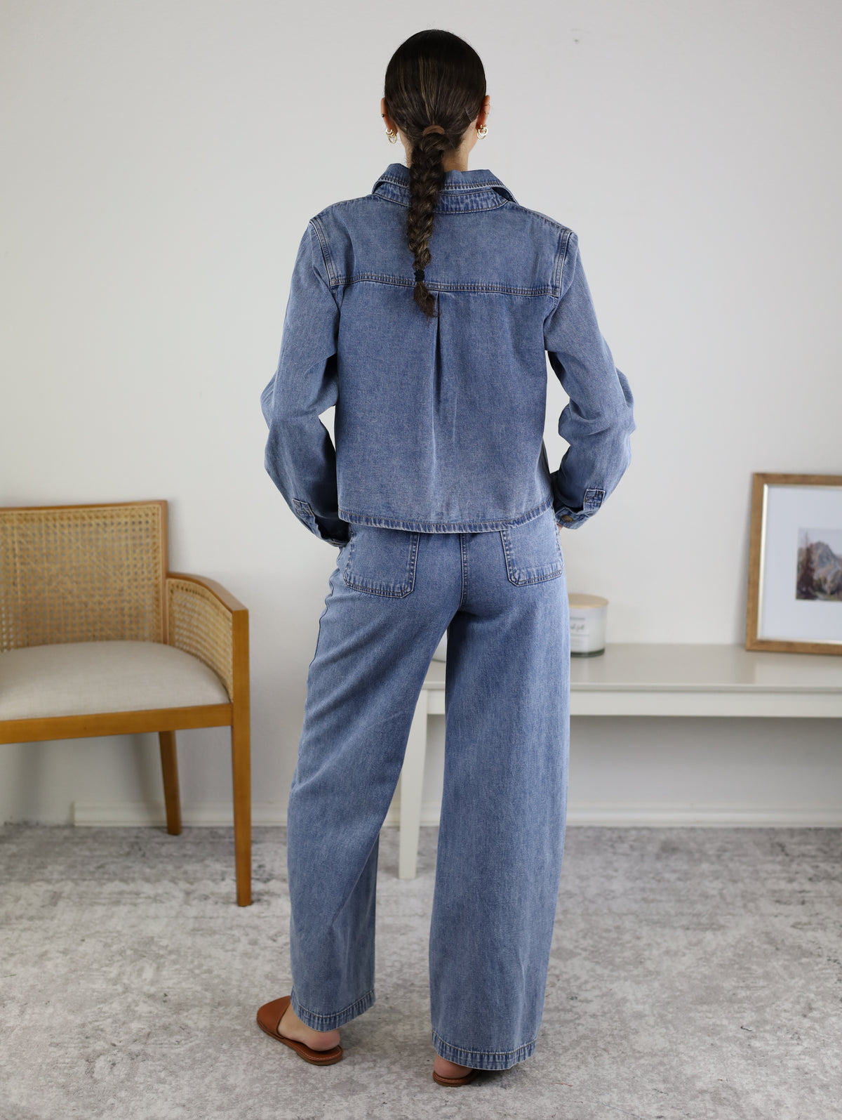 A person with long hair in a ponytail stands facing away, wearing the Adalyn Acid Wash Denim Boxy Shirt paired with wide-leg denim pants. They are indoors near a wooden chair and a white table adorned with decor.