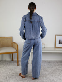 A person with long hair in a ponytail stands facing away, wearing the Adalyn Acid Wash Denim Boxy Shirt paired with wide-leg denim pants. They are indoors near a wooden chair and a white table adorned with decor.