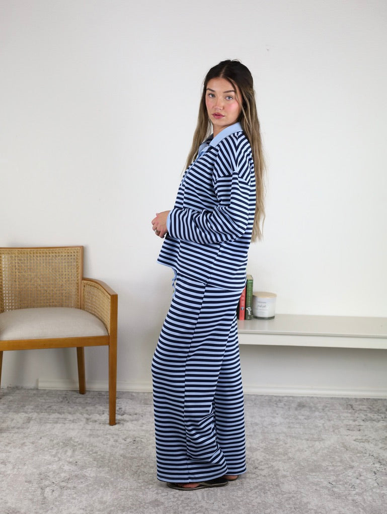 A woman stands sideways indoors, near a wooden chair and a low shelf with books and decor, wearing the Ivy Striped Relaxed Top & Wide Leg Pants Knit Set, which features a blue and white striped long-sleeve top paired with matching wide-leg pants.