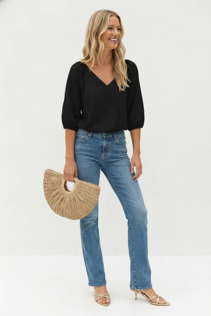 A woman wearing a Naked Zebra - Elastic Puff Sleeve V-neck Blouse: Burgundy / L, blue jeans, and gold heels, holding a straw half-moon clutch.