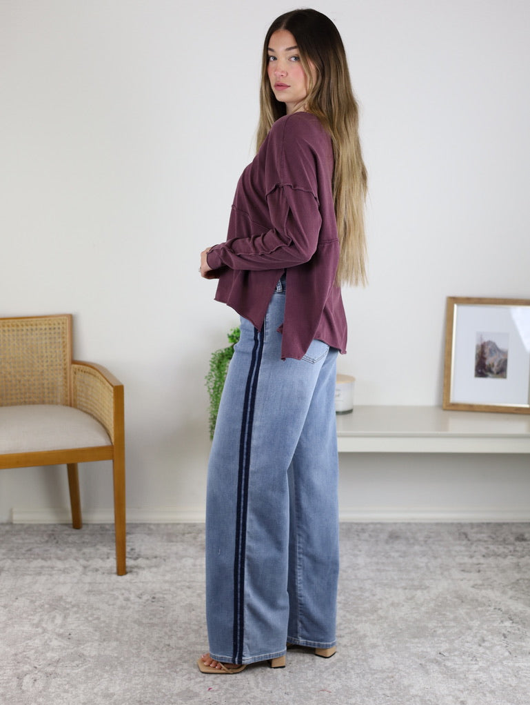 A woman with long hair wears the Ester Raw Seam Henley in maroon and loose blue jeans with a dark side stripe, standing sideways in a room with a chair, framed picture, and plant.