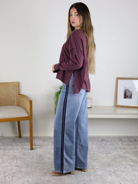 A woman with long hair wears the Ester Raw Seam Henley in maroon and loose blue jeans with a dark side stripe, standing sideways in a room with a chair, framed picture, and plant.
