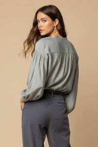 A woman wearing an elegant sage green Naked Zebra - Button-down Blouse With Gathered Neckline with long sleeves and dark grey trousers.