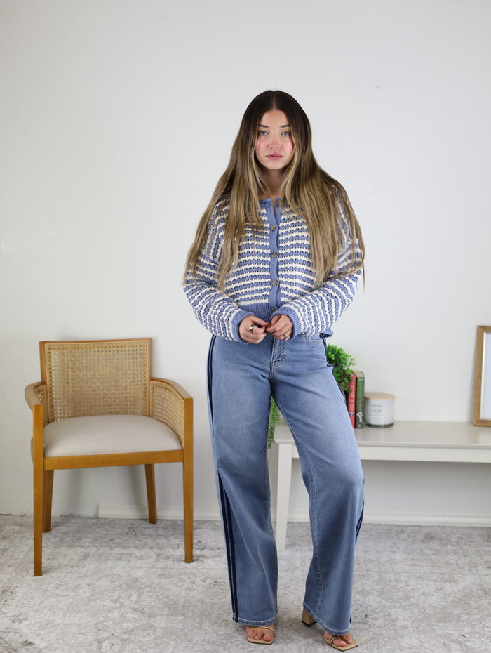 A woman with long hair stands indoors, wearing the Bellamy Textured Striped Crochet Cardigan and wide-leg jeans. She poses beside a wooden chair, with a small table and decor in the background.