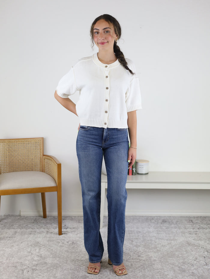 A woman stands indoors wearing the Nathan Raw Edge Short Sleeve Cardigan in white, paired with blue jeans and sandals. She has long brown hair styled in a braid and is positioned next to a wooden chair and a white bench topped with books and a candle.