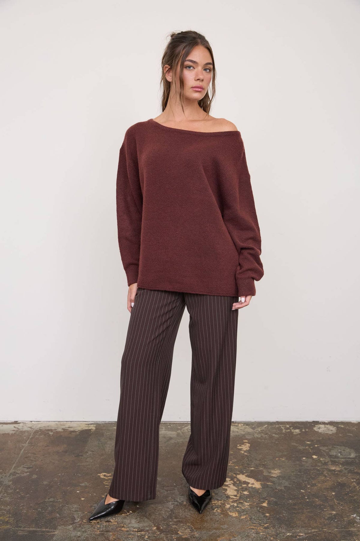 A woman stands on a worn concrete floor against a plain white wall, wearing The Workshop Studio - BOAT NECK OVERSIZED KNIT PULLOVER SWEATER in oatmeal. The loose, oversized knit sweater features a boat neck. She has long brown hair and pairs the sweater with brown pinstripe wide-leg pants and black pointed-toe heels.