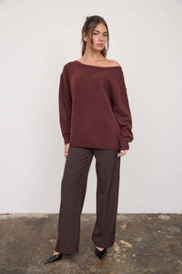 A woman stands on a worn concrete floor against a plain white wall, wearing The Workshop Studio - BOAT NECK OVERSIZED KNIT PULLOVER SWEATER in oatmeal. The loose, oversized knit sweater features a boat neck. She has long brown hair and pairs the sweater with brown pinstripe wide-leg pants and black pointed-toe heels.