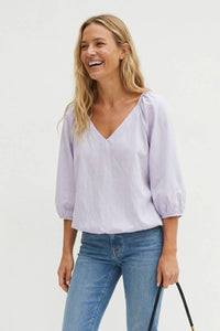 A laughing woman wears a Naked Zebra - Elastic Puff Sleeve V-neck Blouse: Burgundy / L, a light purple V-neck puff sleeve blouse, and blue jeans.