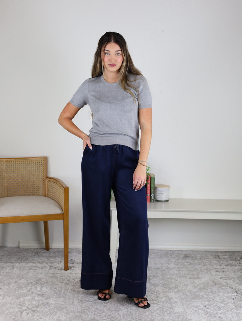 A woman stands indoors wearing the Bellini Basic Sweater Tee in gray, paired with navy wide-leg pants and black sandals. She is positioned near a wooden chair and a white bench adorned with books and a candle. The background is a plain white wall.