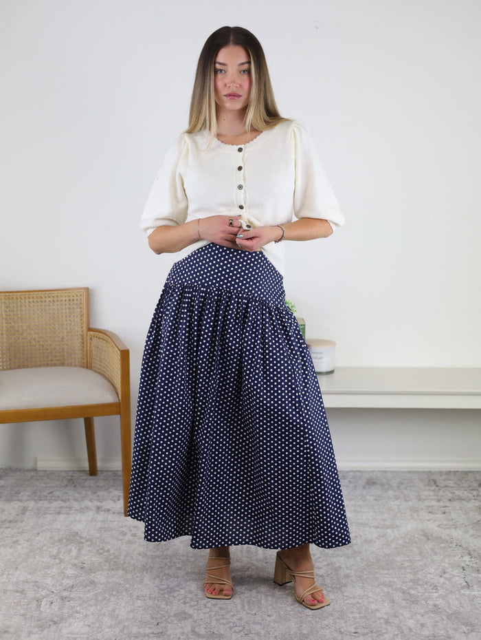 A woman stands indoors wearing a white short-sleeve buttoned top and the Selene Drop Waist Skirt—a long, navy blue 100% cotton midi skirt adorned with white polka dots. She has light brown hair and is wearing sandals. In the background, there’s a wooden chair and a bench.