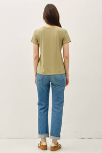 A woman with long brown hair stands facing away, wearing the Fern Basic Bamboo Tee in a relaxed olive green fit, paired with blue jeans and tan sneakers against a plain light background.
