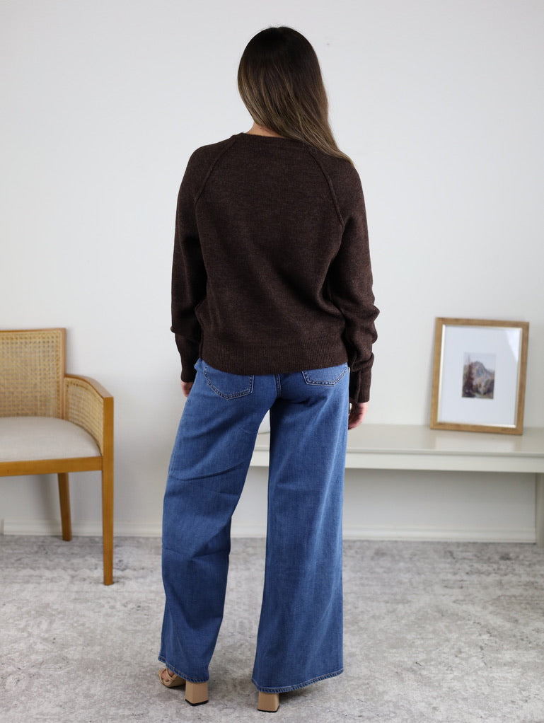 A woman stands facing away, wearing the relaxed fit Theo Sweater and wide-leg blue jeans in a room with a chair, a framed picture on a bench, and a light-colored carpet.