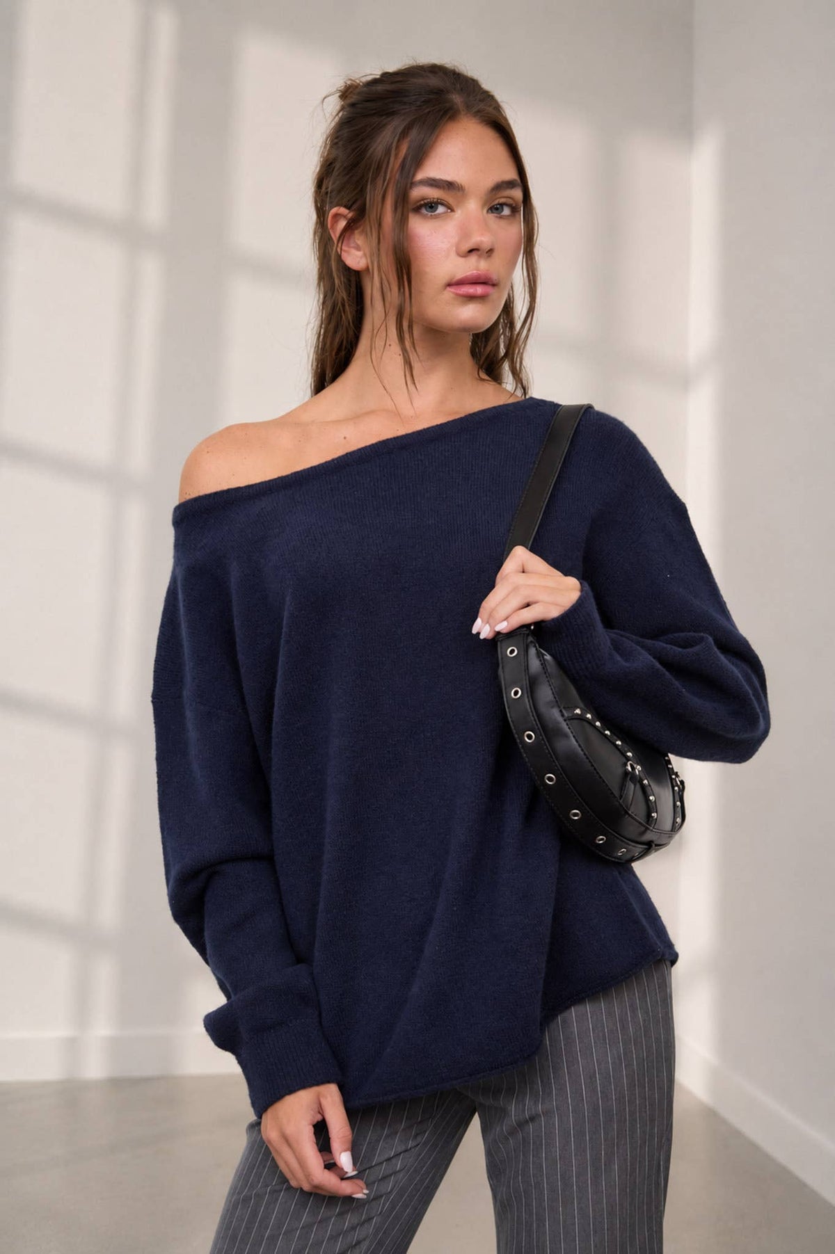 A woman stands in a sunlit room with white walls and large windows, wearing The Workshop Studio’s Boat Neck Oversized Knit Pullover Sweater in oatmeal styled with pinstriped pants and a black crossbody bag.