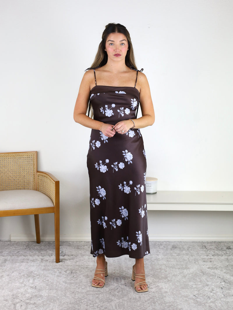 A woman stands indoors wearing the Robbie Spaghetti Strap Floral Midi Dress, featuring a delicate brown floral print and slim straps. She pairs it with light-colored heels, facing forward as a wooden chair and a white bench sit on a light carpet in the background.