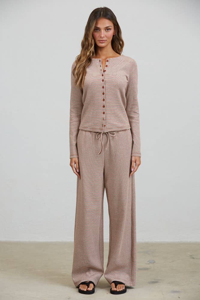 A woman stands against a plain background wearing the Creamon CTP3592 BROWN Lounge Striped Button Down Top & Pants Set in brown stripe, featuring a beige long sleeve button down top and wide leg pants, paired with black sandals.