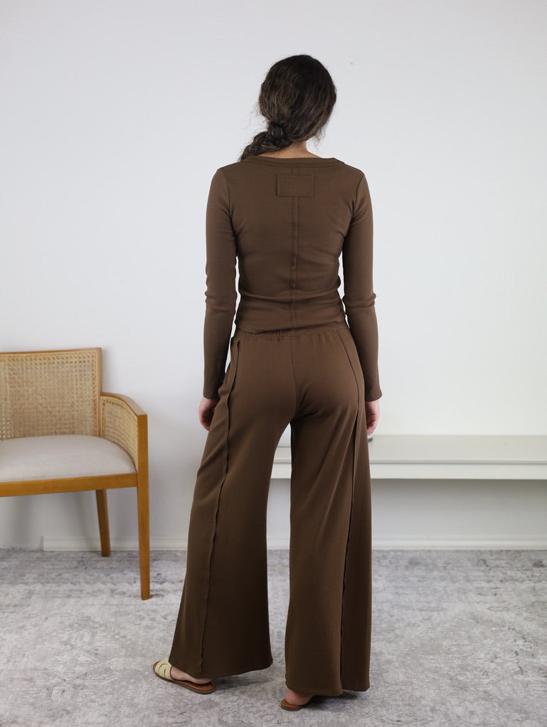 A person with long hair, dressed in the Statler Lounge Set—a brown henley top and wide-leg pants—stands facing away in a neutral indoor space with a gray carpet. On the left is a wooden chair featuring a woven back.