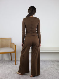 A person with long hair, dressed in the Statler Lounge Set—a brown henley top and wide-leg pants—stands facing away in a neutral indoor space with a gray carpet. On the left is a wooden chair featuring a woven back.