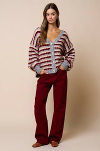 A woman stands with her hands in her pockets, wearing the Lalavon Relaxed Stripe V-Neck Button Cardigan in GREY-BURGUNDY-BEIGE (size L), which features large buttons, a loose fit, and bold gray, white, and red stripes. She pairs it with burgundy pants and brown shoes against a plain beige background.