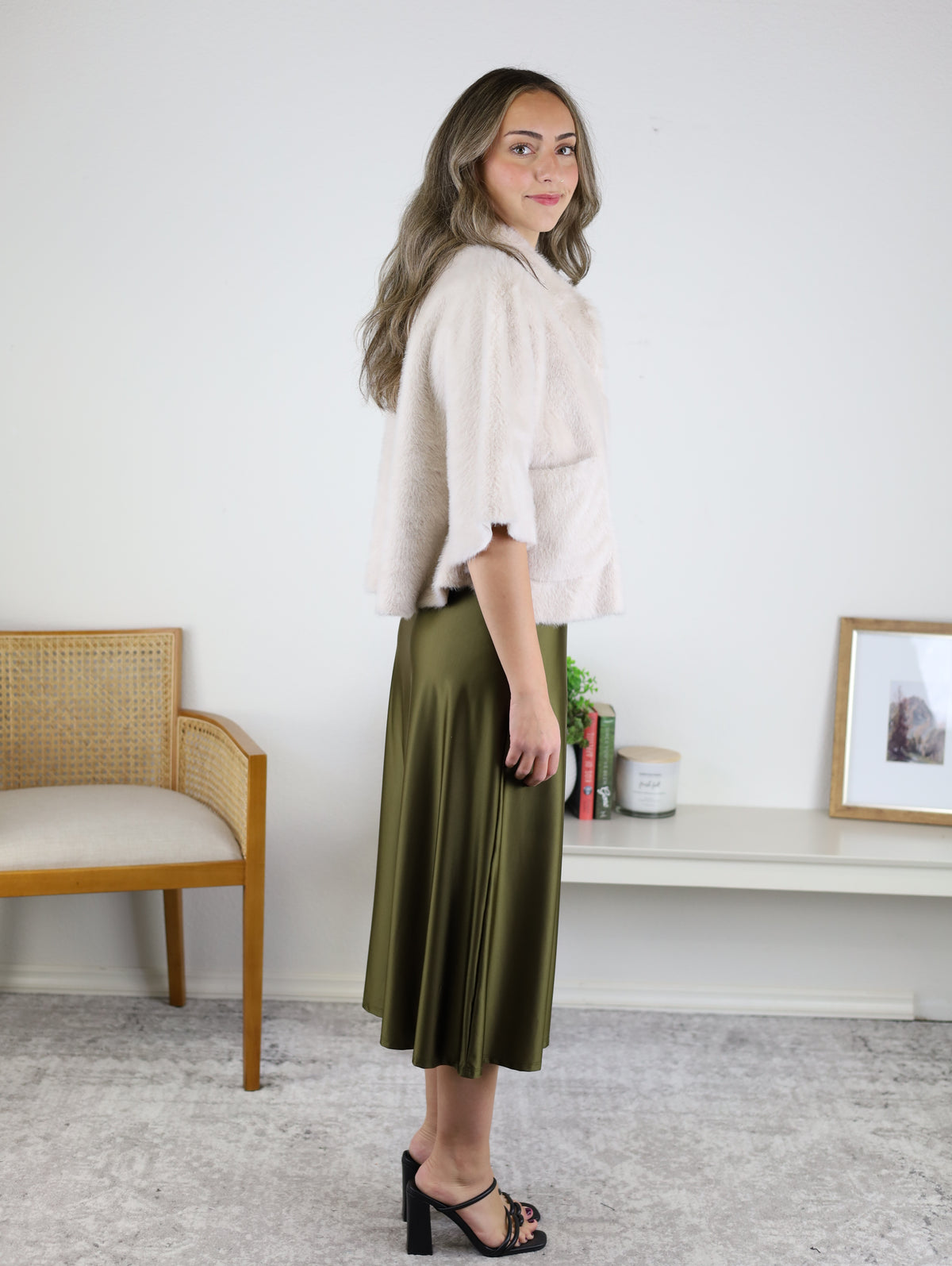 A person wearing a light-colored, cropped Jordan Fur Cape Sleeve Jacket and an olive green midi skirt, paired with black heeled sandals.
