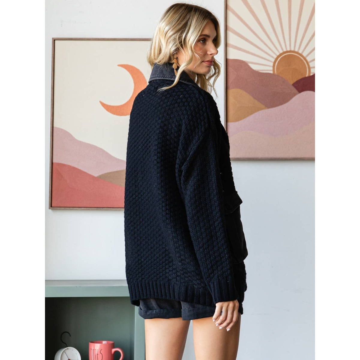 A woman with long blonde hair wears the Veveret - Knit Body Denim Layerd Jacket: Cream / Denim / L, standing sideways before abstract sun and moon artwork over colorful hills.