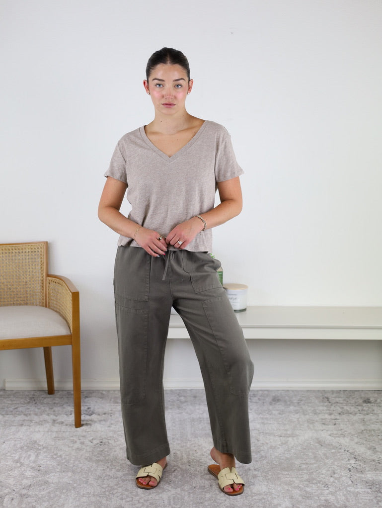 A woman stands indoors wearing the Nomad V Neck Tee in beige, crafted from soft knit fabric, paired with olive green drawstring pants and cream sandals. She stands on a light rug next to a wooden chair and a white shelf against a plain wall.