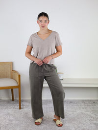 A woman stands indoors wearing the Nomad V Neck Tee in beige, crafted from soft knit fabric, paired with olive green drawstring pants and cream sandals. She stands on a light rug next to a wooden chair and a white shelf against a plain wall.