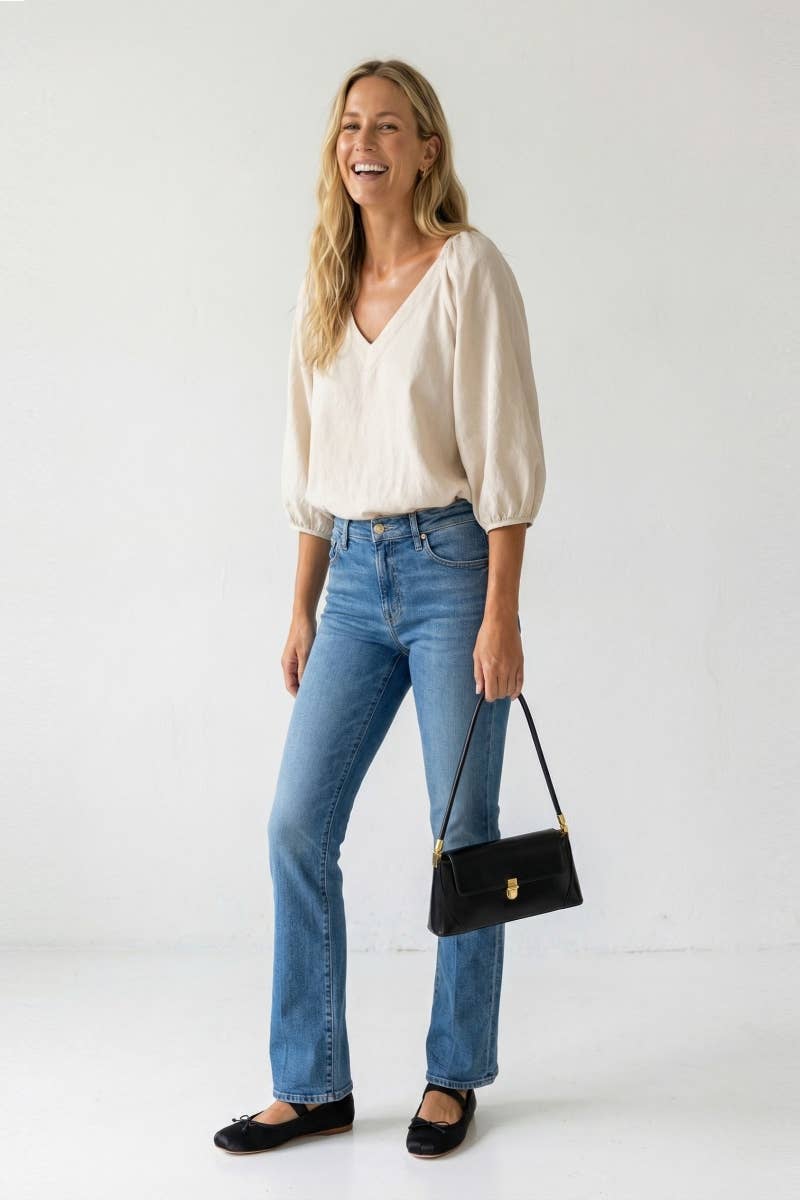 A woman smiles while wearing a Naked Zebra - Elastic Puff Sleeve V-neck Blouse: Burgundy / L, blue jeans, and black ballet flats, carrying a small black shoulder bag.