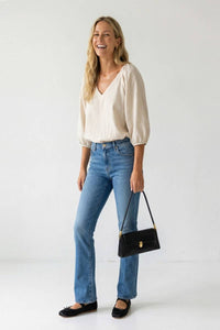 A woman smiles while wearing a Naked Zebra - Elastic Puff Sleeve V-neck Blouse: Burgundy / L, blue jeans, and black ballet flats, carrying a small black shoulder bag.