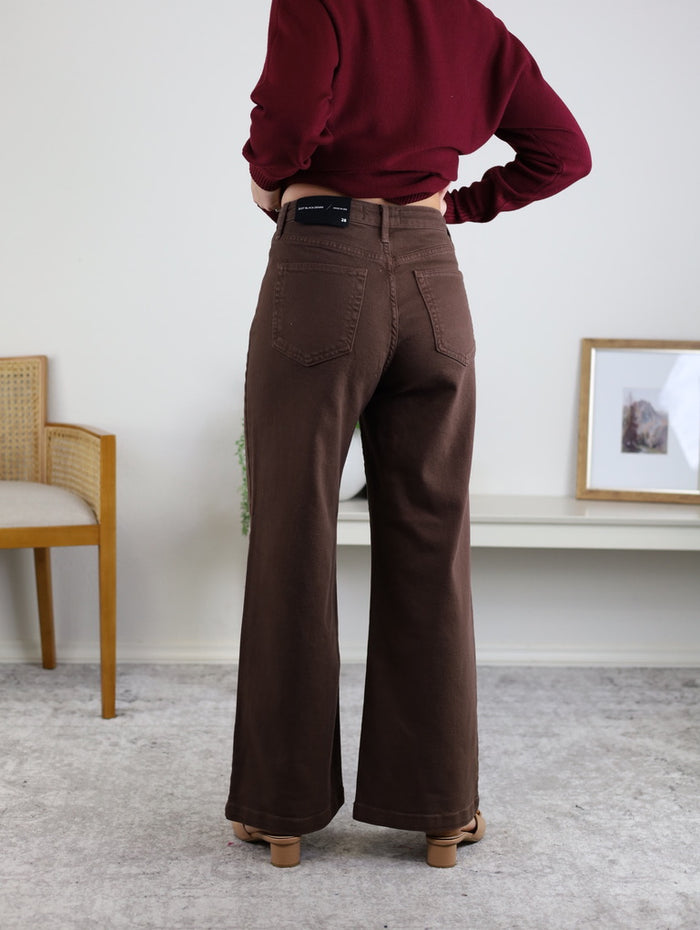 A person wears the Aggie Patch Pocket Wide Leg jeans and a maroon top, standing indoors with their back to the camera. A chair and framed picture appear in the background.