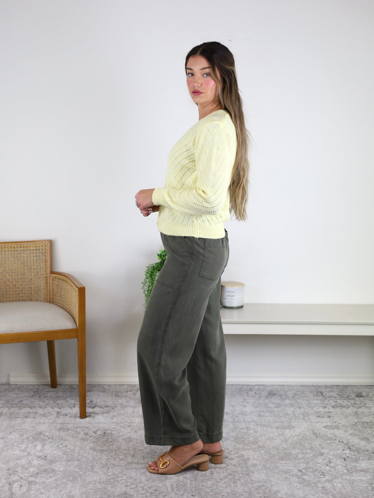 A woman with long hair stands sideways indoors, wearing a light yellow sweater, tan sandals, and the Marbella Wide Leg Tencel Pant in olive green, featuring a breathable flowy design with a drawstring elastic waist. She is on a gray carpet next to a wooden chair and a white bench adorned with a potted plant and candle.
