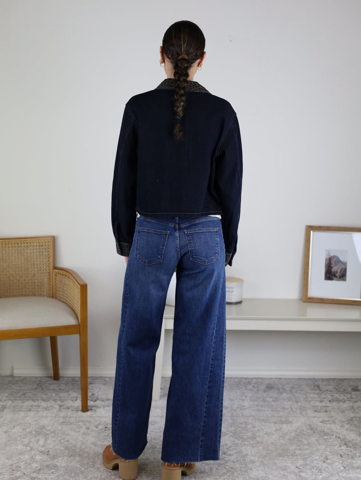 A person with a braid stands facing away, wearing the Autumn Leopard Collar Denim Shacket, wide-leg blue jeans, and tan shoes in a neutral room with a chair, white table, and a framed photo.
