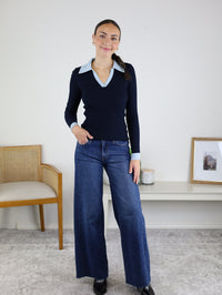 A woman stands indoors, smiling, wearing the Deepsea Ribbed Collared Knit Top with a light blue collar and wide-leg jeans. She poses beside a wooden chair and a white table adorned with a picture frame and cup.