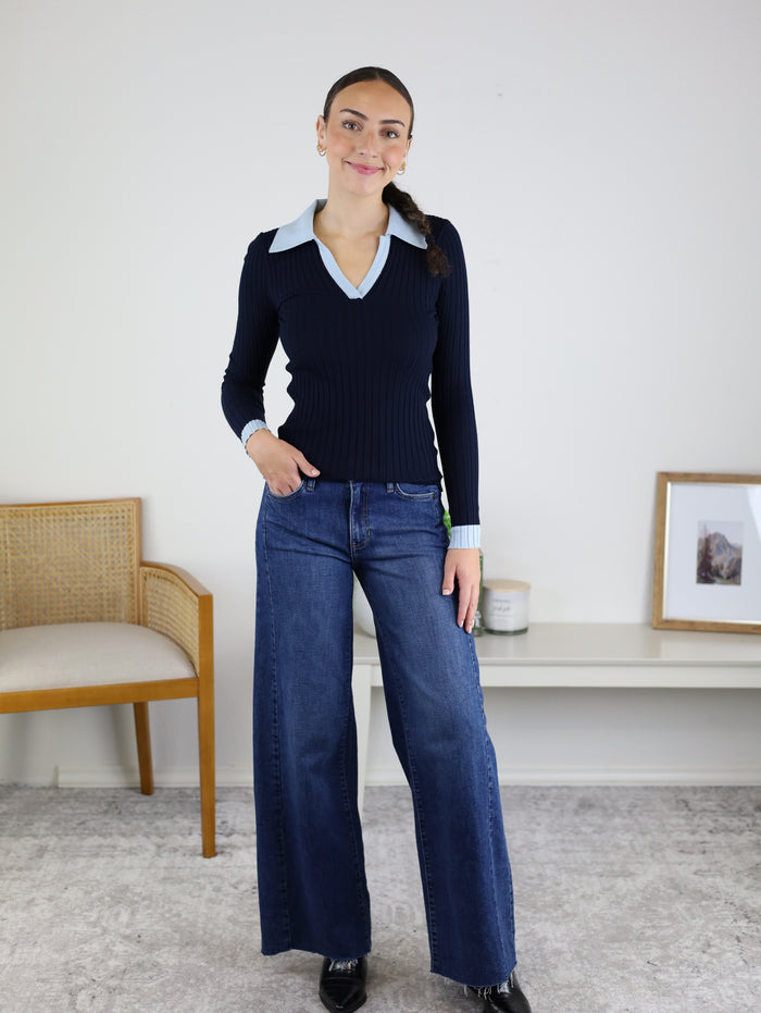 A woman stands indoors, smiling, wearing the Deepsea Ribbed Collared Knit Top with a light blue collar and wide-leg jeans. She poses beside a wooden chair and a white table adorned with a picture frame and cup.