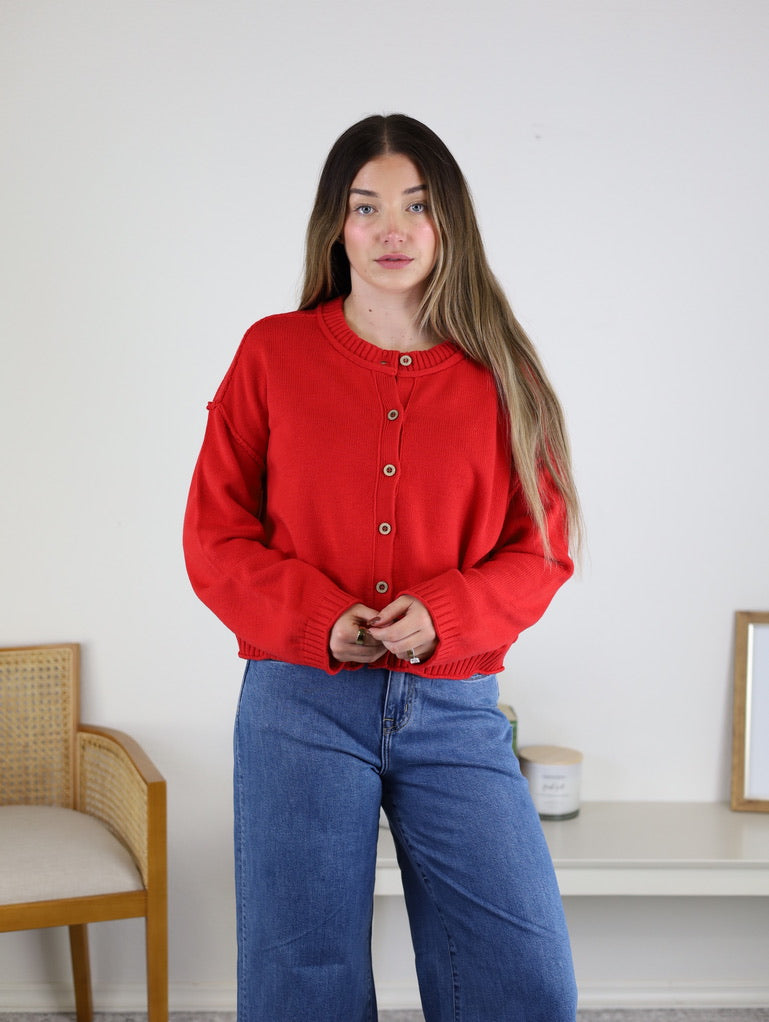 A person wears a red Jude Raw Edge Cardigan, a button-front lightweight knit with raw-edge shoulder detail, paired with blue wide-leg jeans.