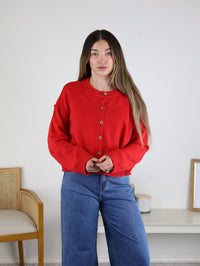 A person wears a red Jude Raw Edge Cardigan, a button-front lightweight knit with raw-edge shoulder detail, paired with blue wide-leg jeans.
