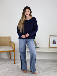 A woman with long hair stands indoors wearing the Bell Contrast Boat Neck Sweater, light blue wide-leg jeans, and sandals beside a table with a framed picture, books, and a chair.
