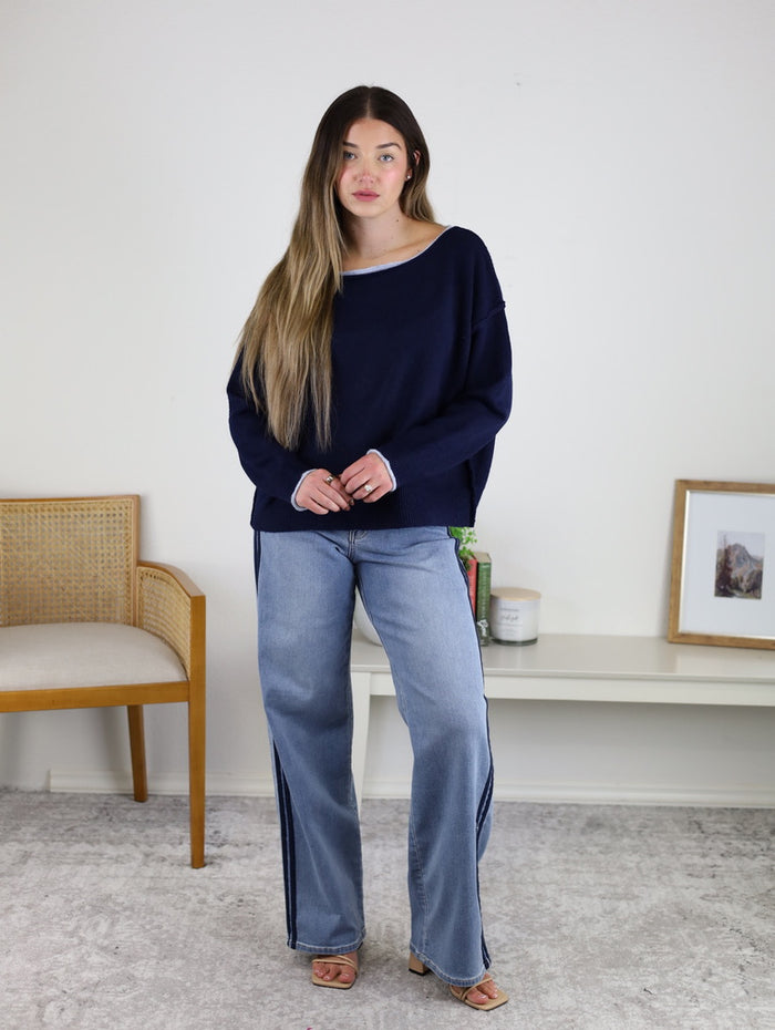 A woman with long hair stands indoors wearing the Bell Contrast Boat Neck Sweater, light blue wide-leg jeans, and sandals beside a table with a framed picture, books, and a chair.